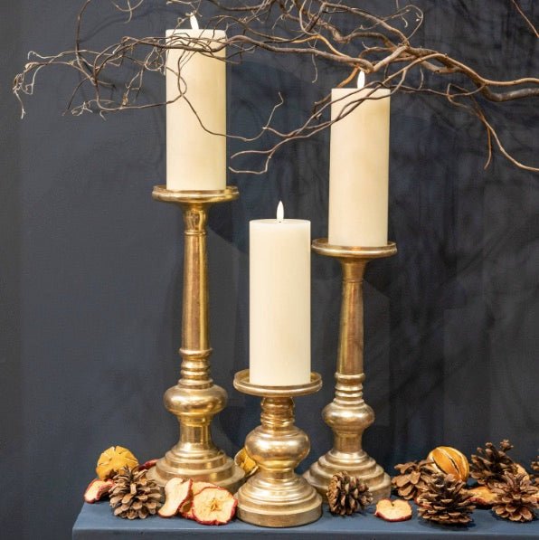 display of 3 LED candles on aged brass effect candlesticks of varying heights, displayed with dried branches, pine cones and dried fruits