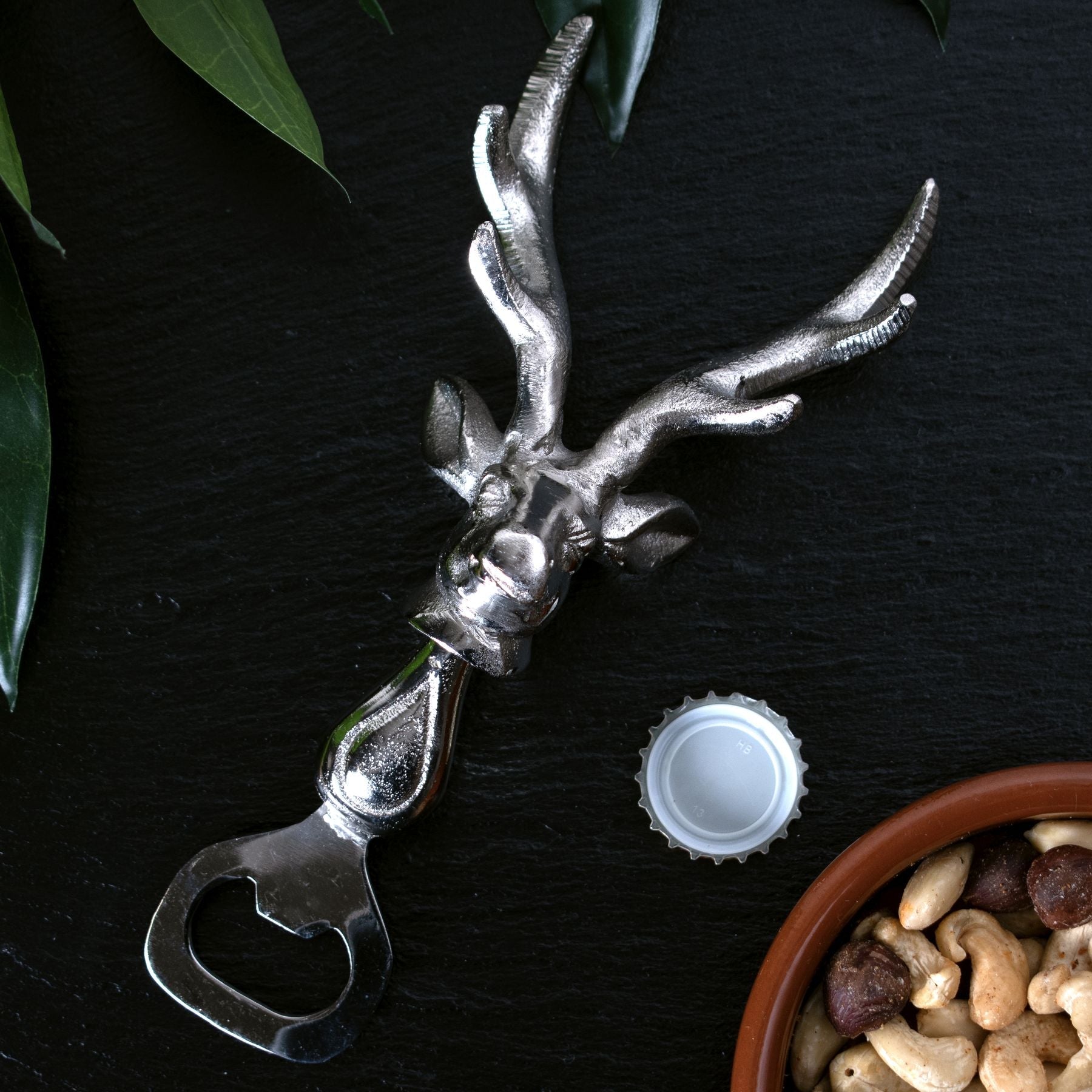 silver stag head bottle opener laid down next to bottle top and small bowl of mixed nuts