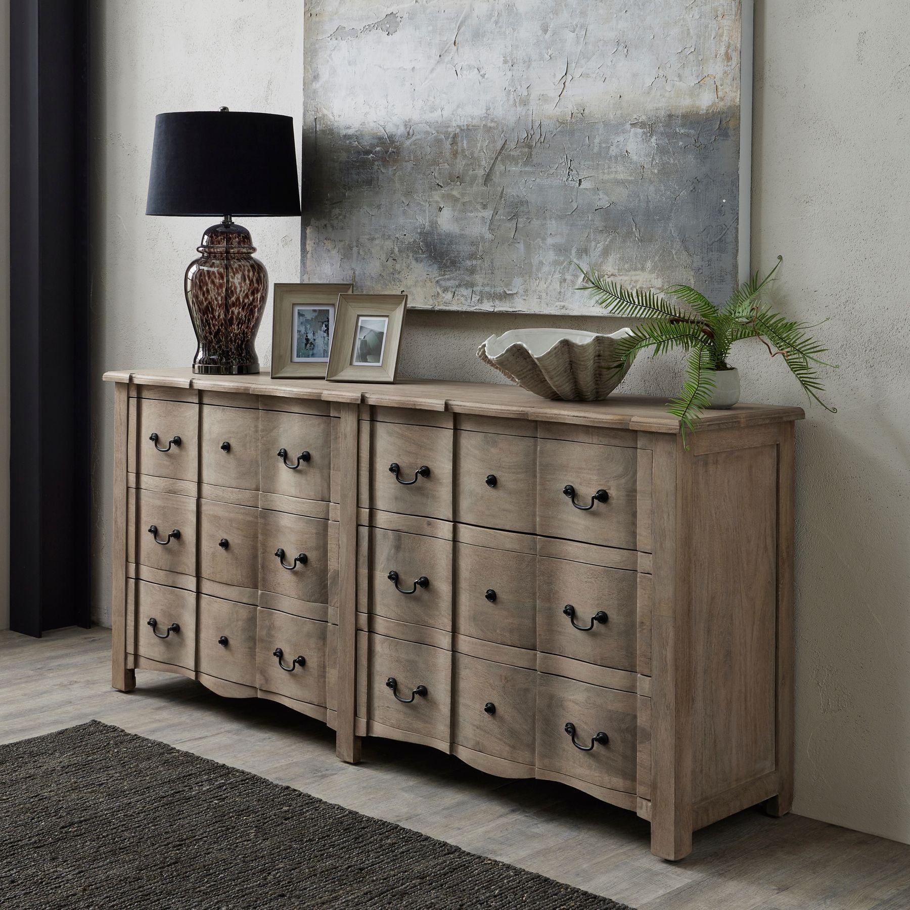 chest of drawers against wall, with art work above. Lamp, two photo frames, shell bowl and artificial fern to the top