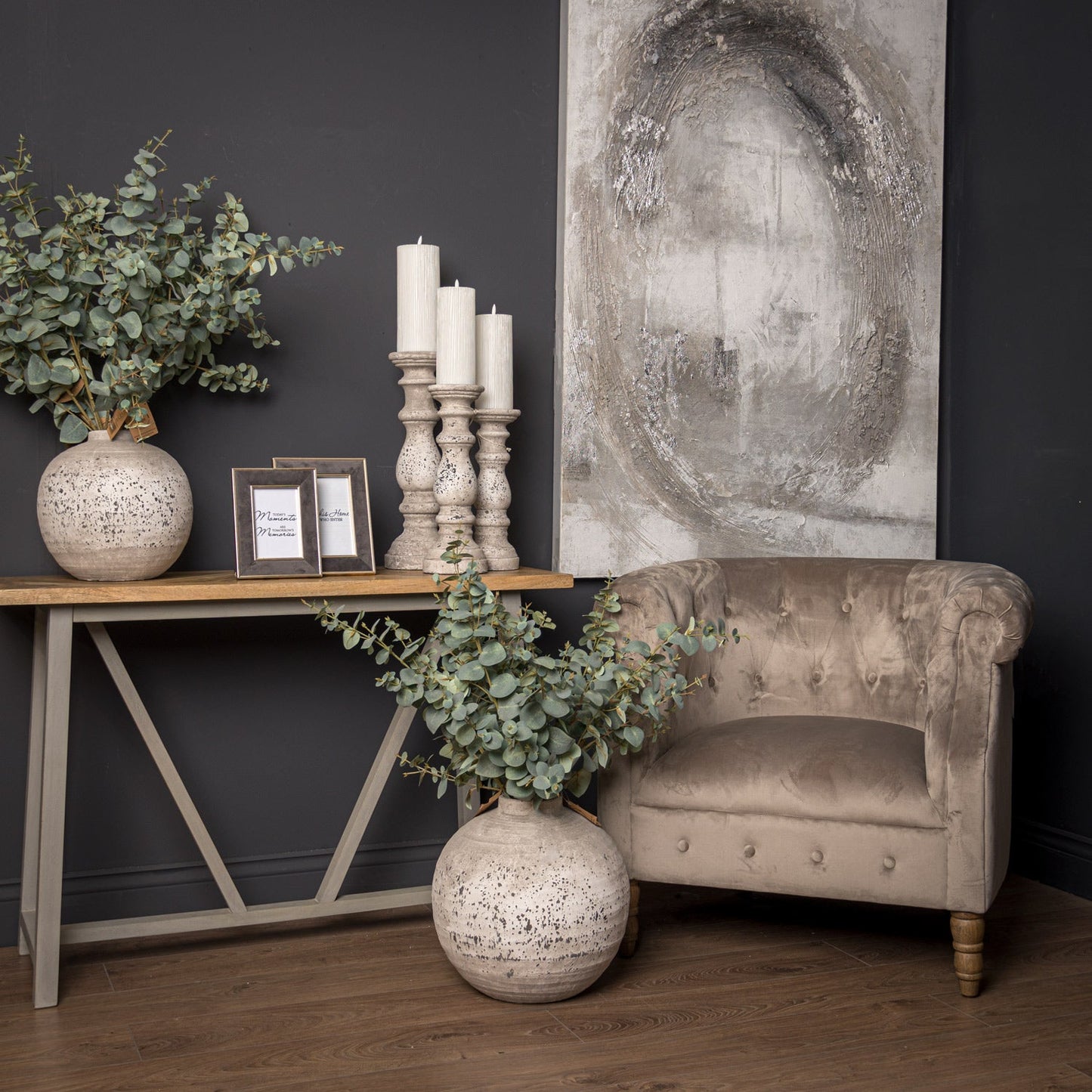 lifestyle image with small Aged Stone globe vase on top of console table with artificial eucalyptus in, grey velvet photo frames next to it and all three sizes of aged stone candle holders with ribbed LED candles to the right. Large Aged stone globe vase on the floor with artificial eucalyptus in. Occasional chair behind and grey abstract wall art to the wall behind