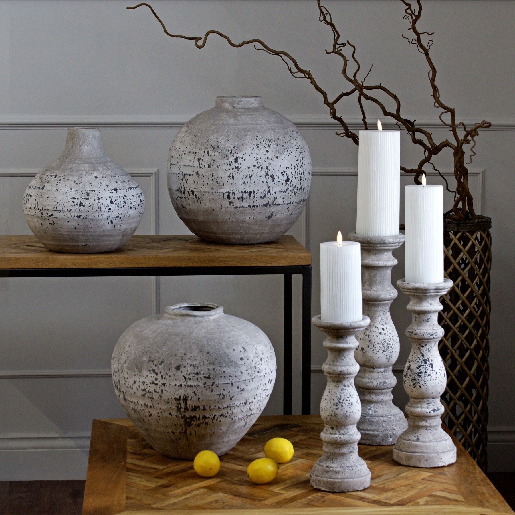 small aged stone globe vase on console table next to aged stone amphora vase. Regal aged stone vase on coffee table in front next to all three sizes of aged stone candle holder, each with ribbed LED candle on. Dried branches can be seen to the background