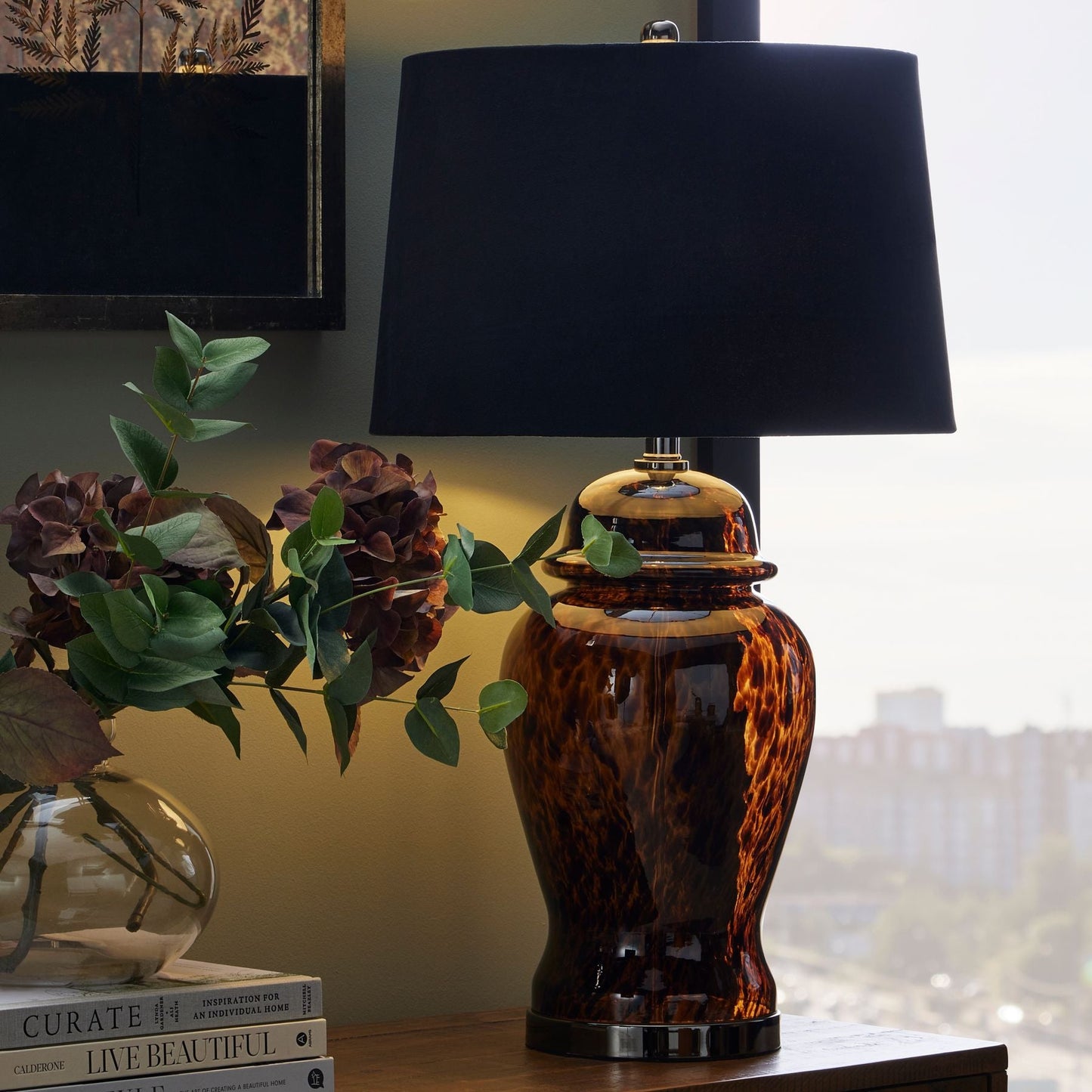 lifestyle image of amber alster lamp on the end of sideboard. Decorative books and chocolate coloured artificial flowers are next to it