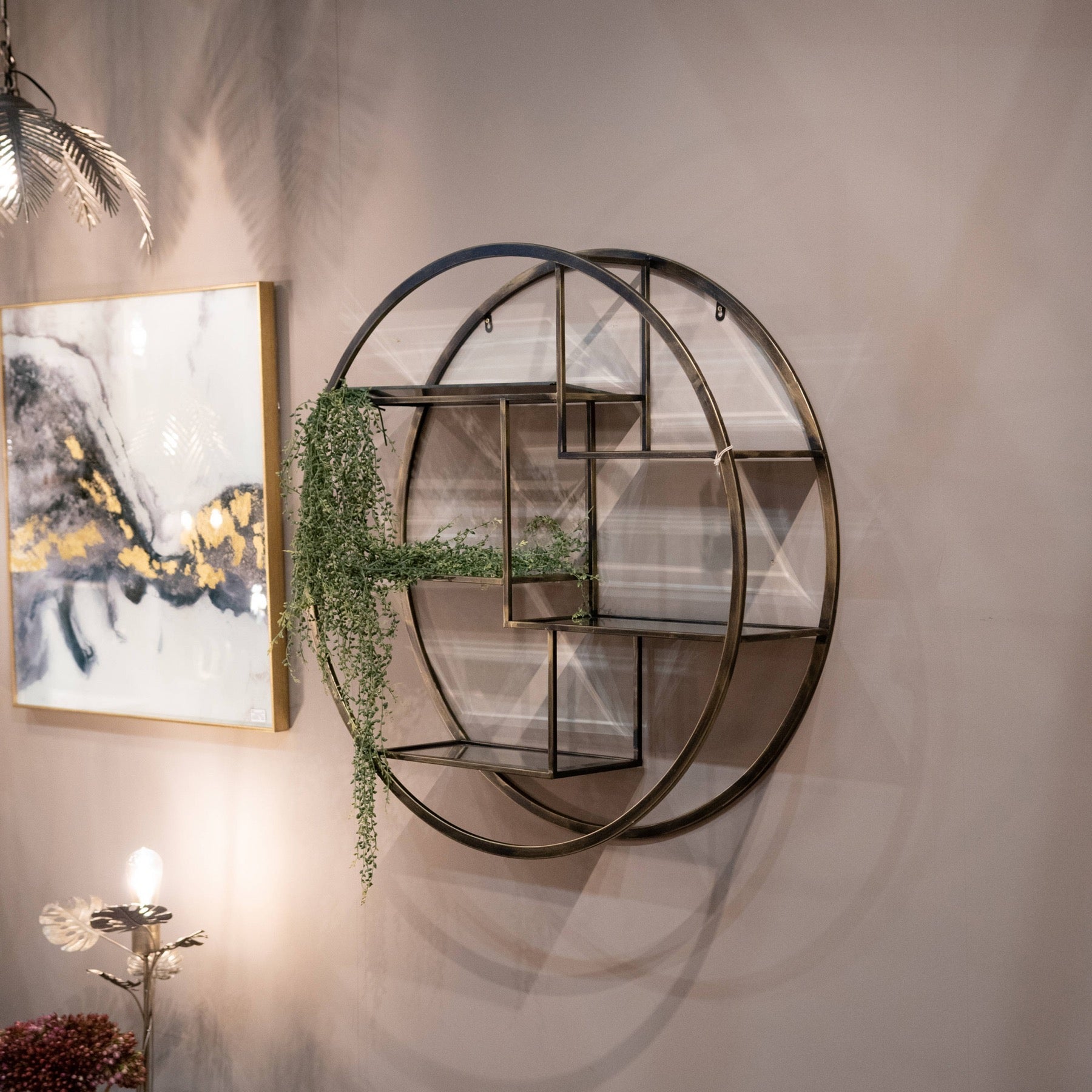 lifestyle image of round antique brass multi shelf unit, an artificial plant sits on the shelf and abstract art is on the wall next to it