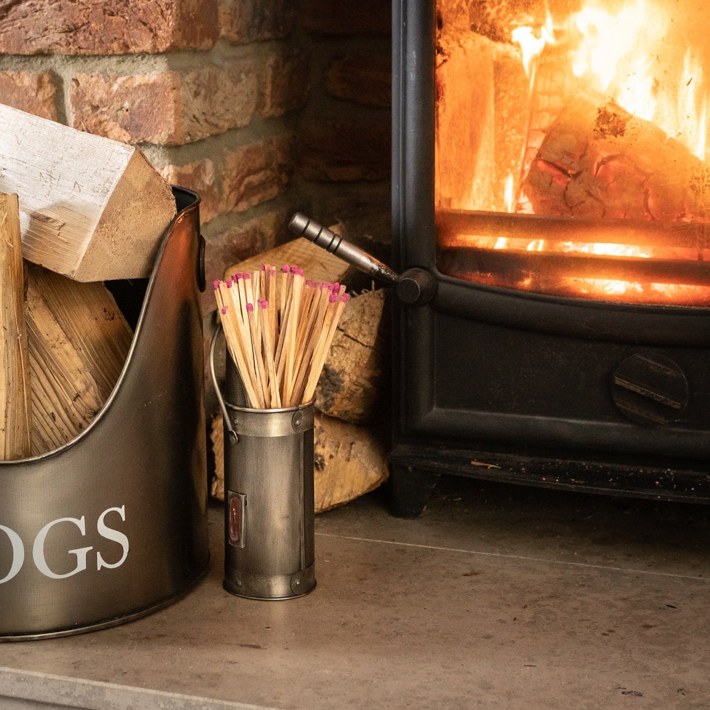 lifestyle image the antique pewter match holder, with matches, sits next to a matching log basket on a stone hearth. A black log burner within an exposed brick fireplace can be seen in the background