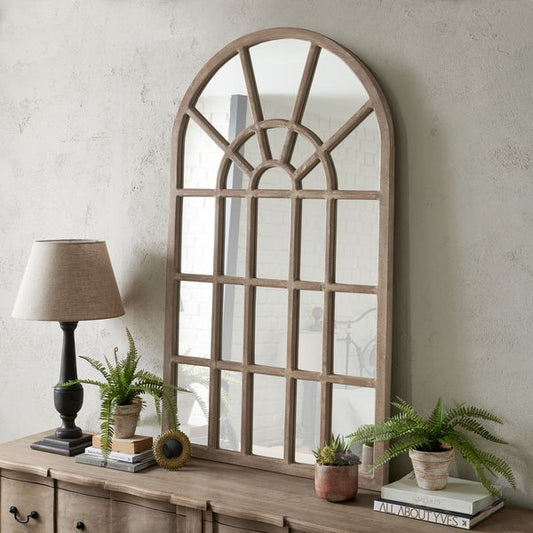 Window Pane style mirror with arched top, leant up against the wall on a sideboard. Sideboard top also has lamp, books and small artificial plants on