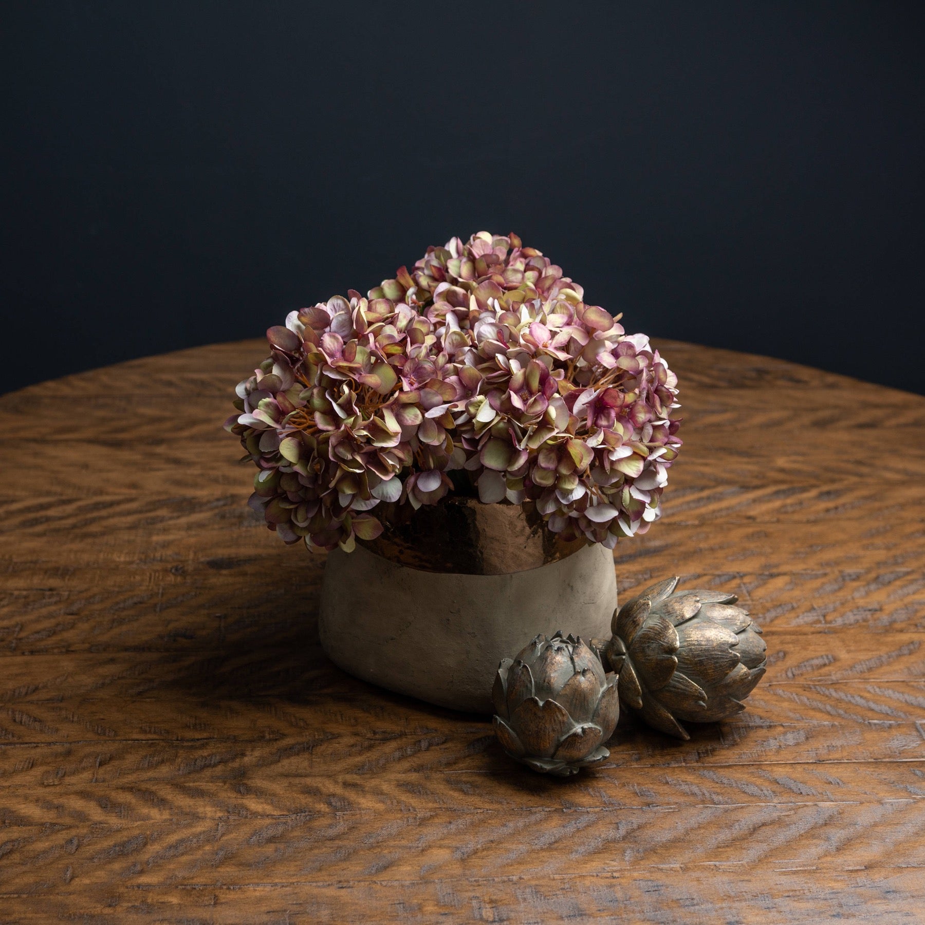 burgundy hydrangeas cut short to fit in short stone vase. Bronze artichoke ornaments are in front of the vase