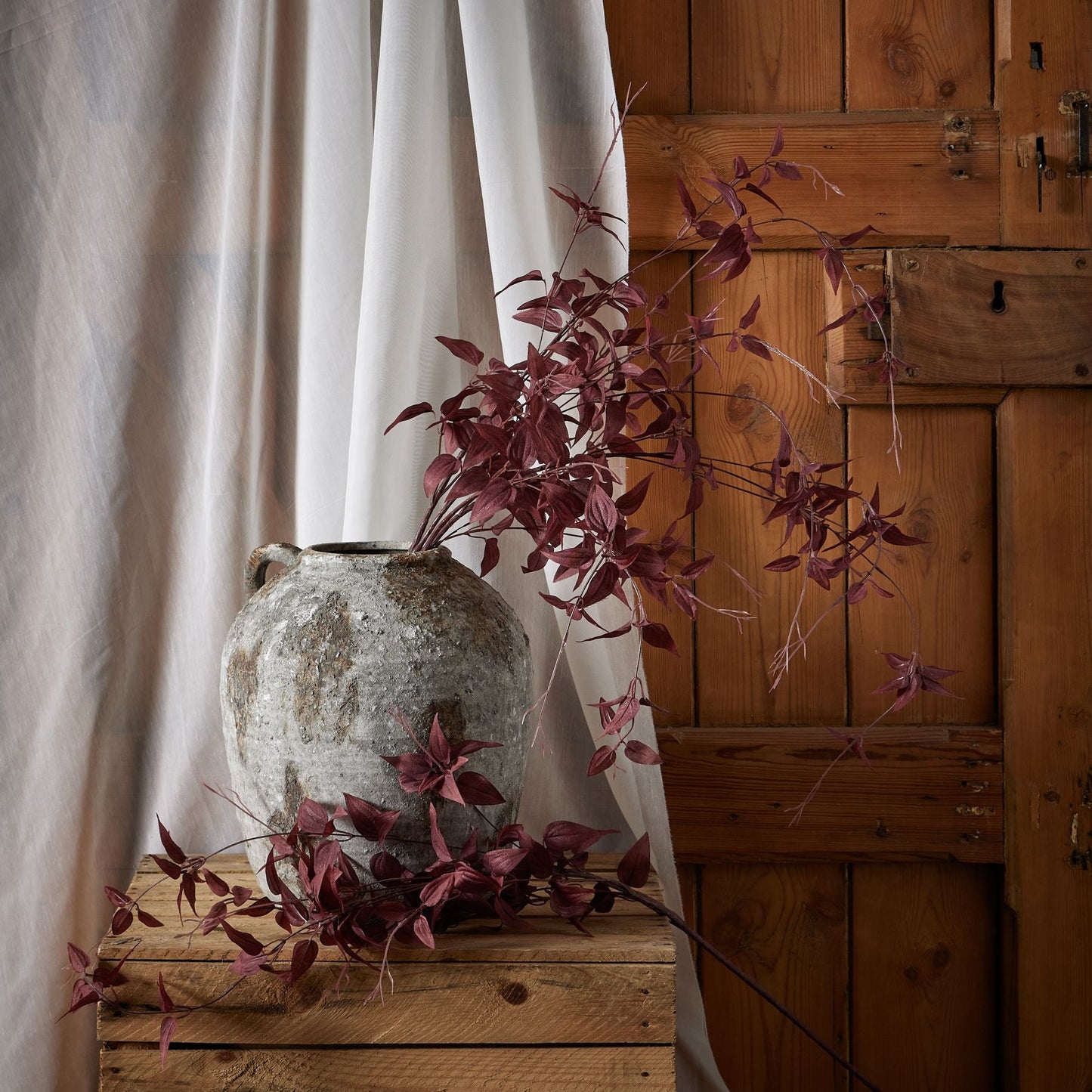 Several stems of burgundy leaf in aged stone vase. Linen hangs behind covering a wooden door