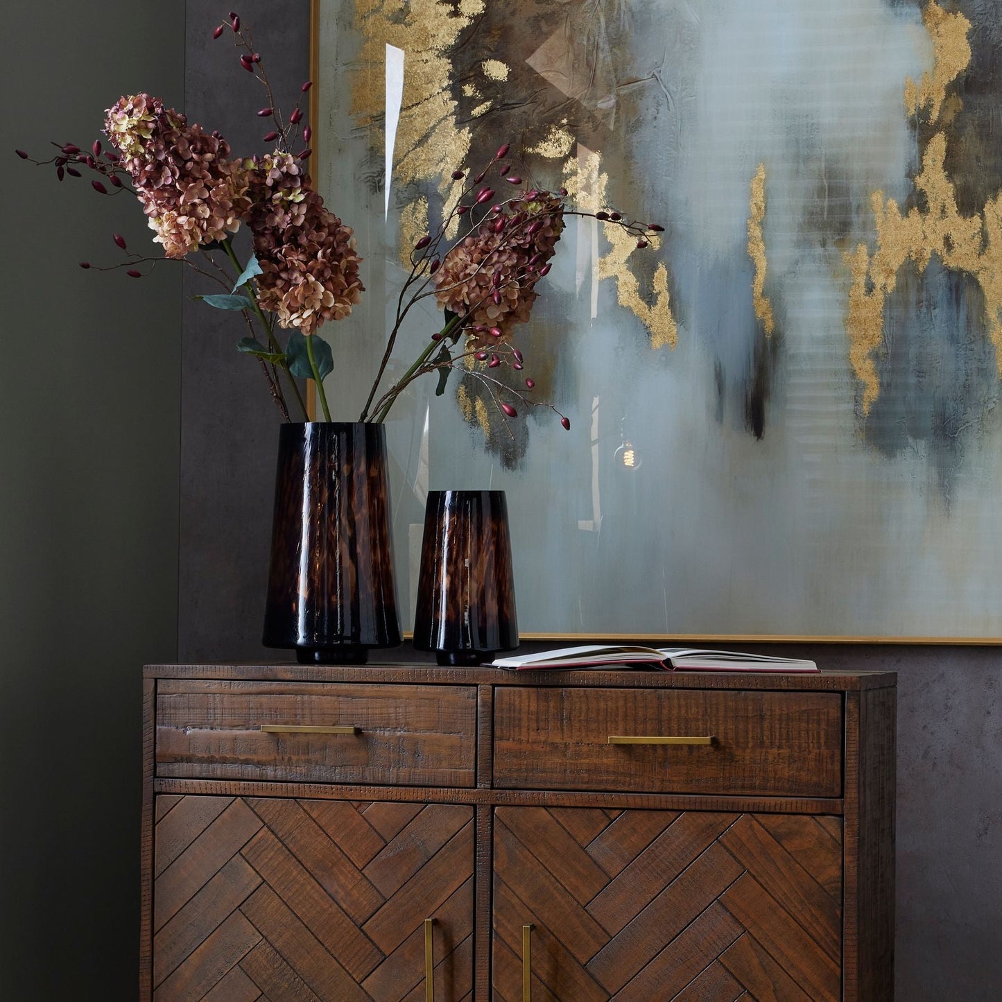 lifestyle image with three spear head hydrangeas in amber dapple tall vase, on top of Bolero 2 door 2 drawer sideboard. An abstract grey and gold picture is on the wall behind