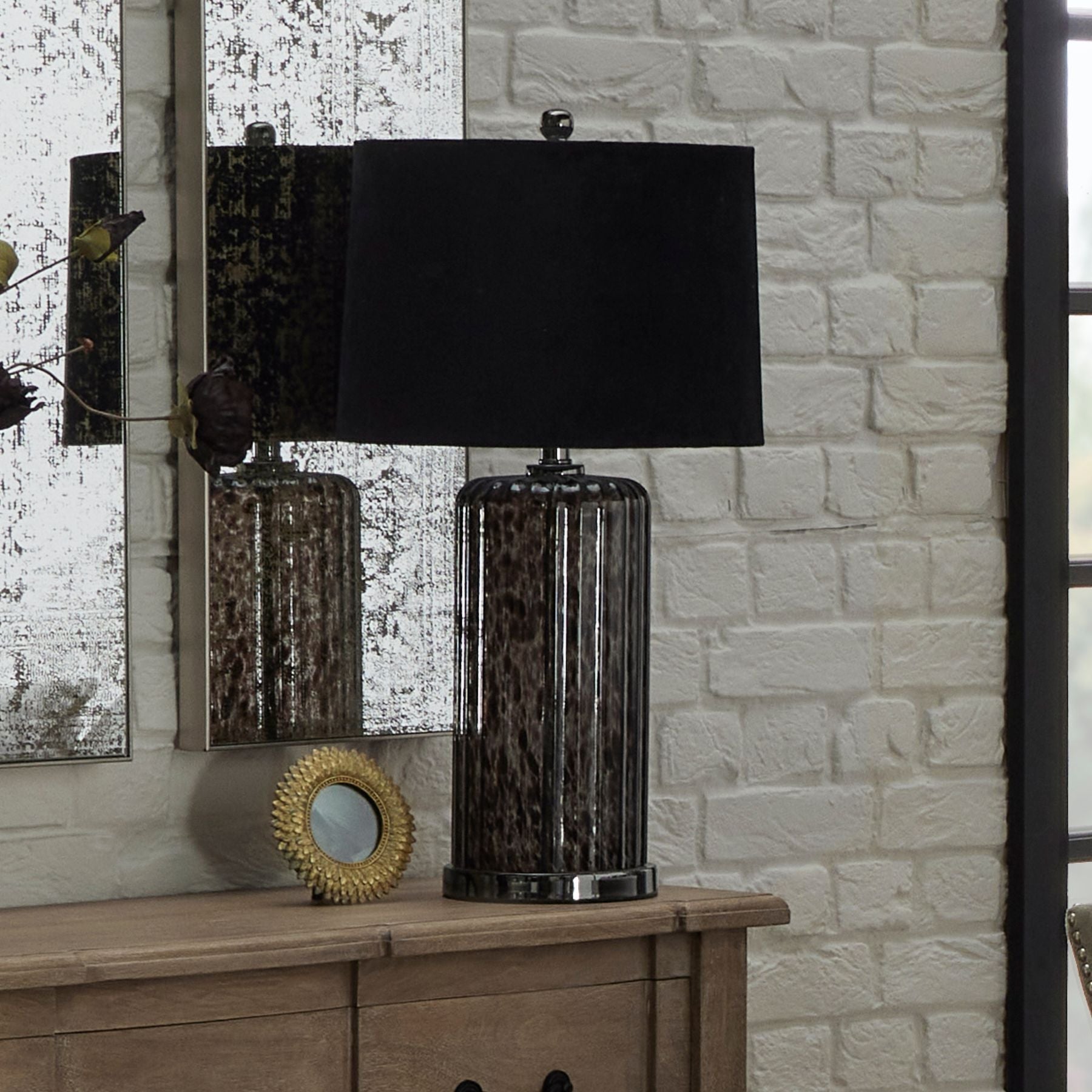 lifestyle image of Calgary table lamp on sideboard, next to round gold feather photo frame and in front of trio of foxed etched mirrors