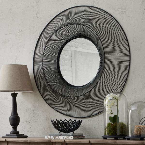 Concave matte black wire mirror above sideboard. On the sideboard is a lamp to the left, books with fruit bowl on in the centre, and then two cloches with artificial orchids in to the right