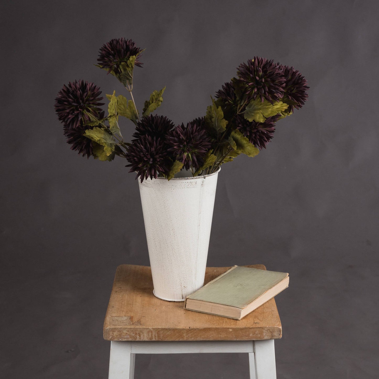 several stems of chocolate brown chrysanthemum flowers in distressed white bucket on top of wooden bench with antique style book next to them