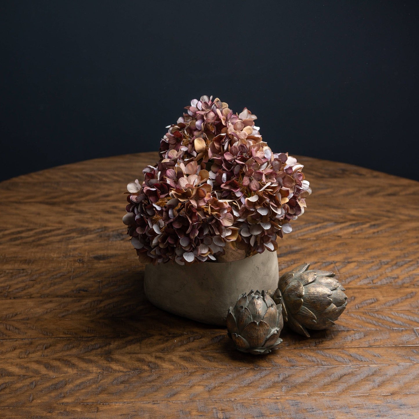 Coffee hydrangeas cut down in short stone vase. Two bronze artichokes are placed in front of the vase