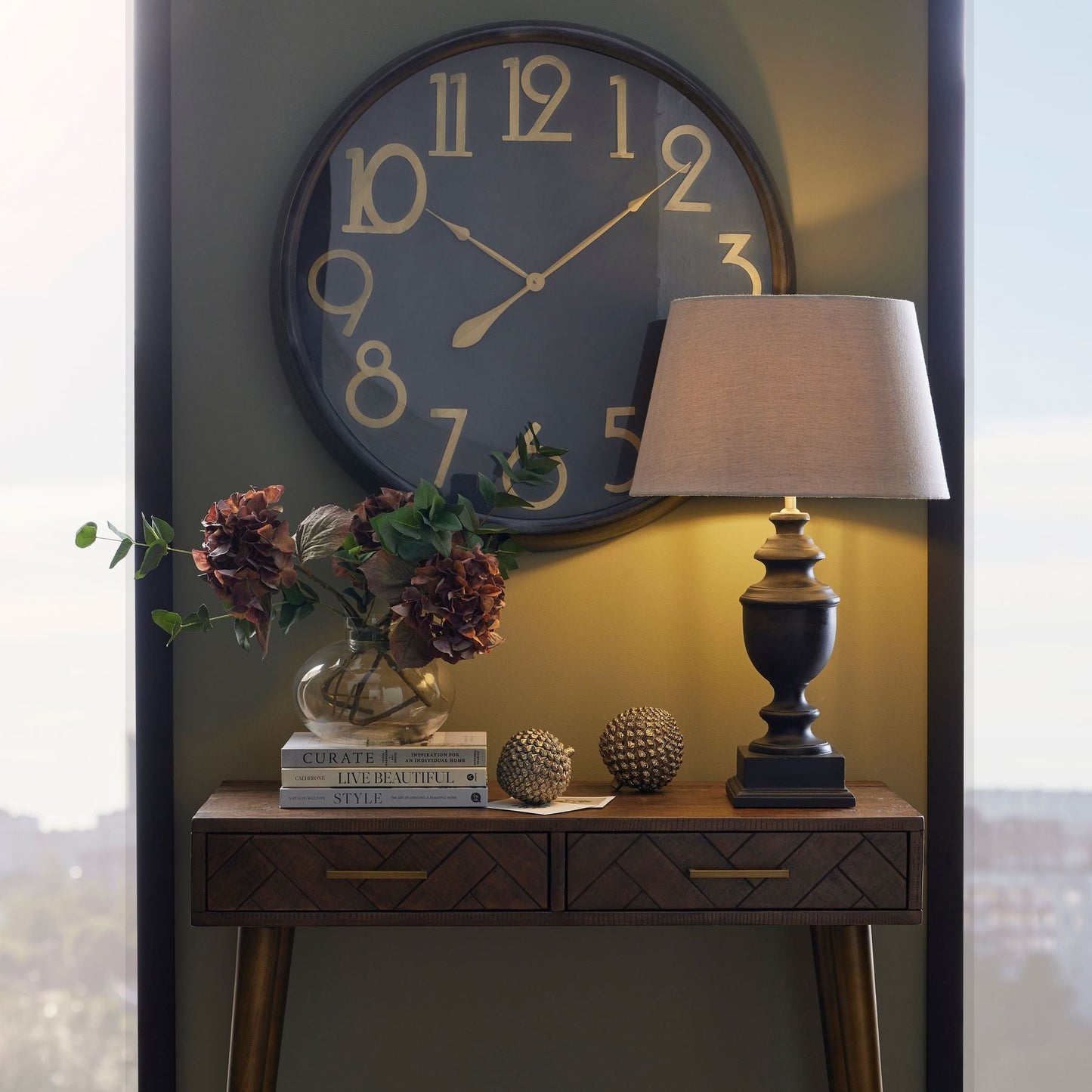 Gatsby black and brass clock above console table. On the console table is wooden lamp with linen shade, two decorative pine cones, decorative books with glass vase and autumnal artificial flowers in