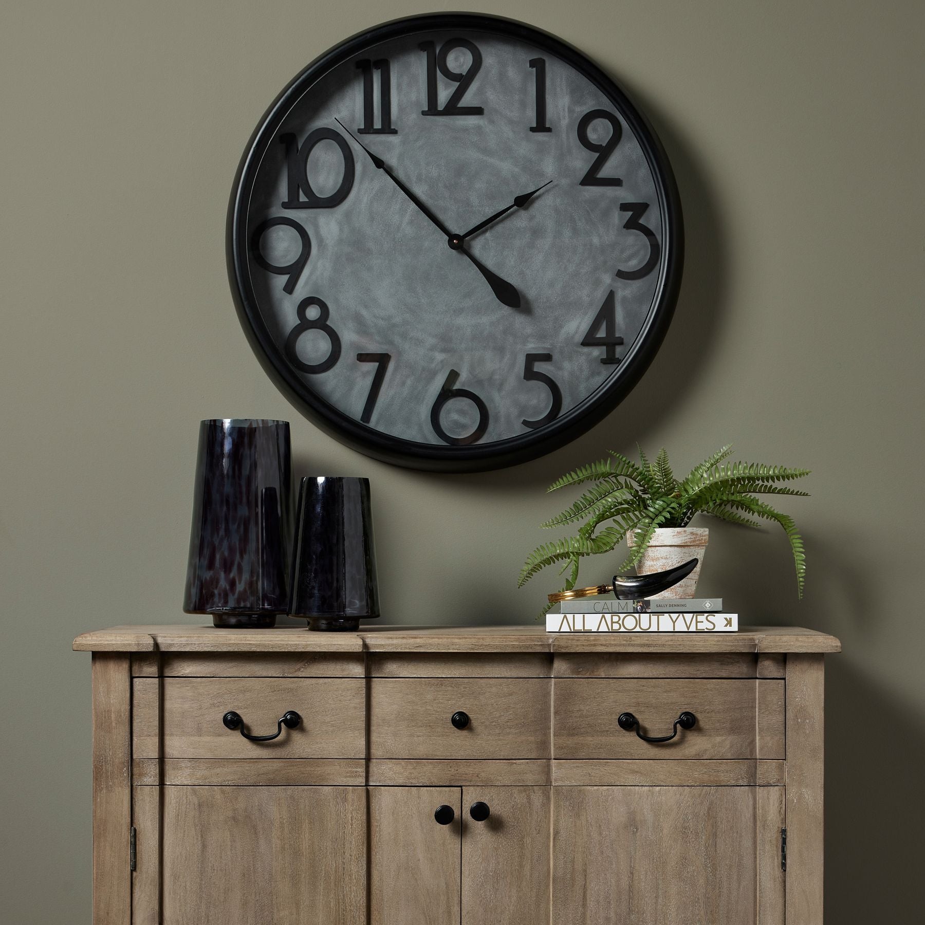 Large clock above 1 drawer 2 door sideboard. Sideboard has two dapple print vases to the left hand side and decorative books to the right with a magnifying glass and artificial fern plant to the top