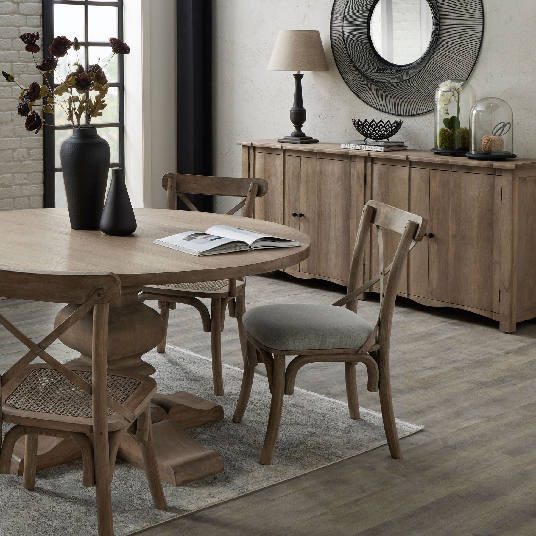 round dining table and chairs in the foreground with large sideboard in the background, which has lamp, bowl and both size cloches on and a large black mirror to the wall behind