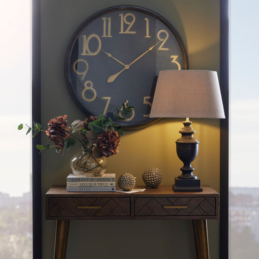 lifestyle image of grey urn lamp with linen shade on 2 drawer console table, two decorative pine cones and artificial flowers are also on the table. A black and brass clocks is mounted on the olive green wall behind