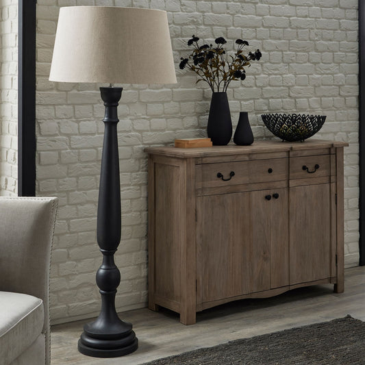 lifestyle image of the Grey wooden floor lamp with linen shade in front of a white painted exposed brick wall, next to a cotswold 2 door 2 drawer sideboard. Two matte black vases and a black decorative bowl sit on top of the sideboard