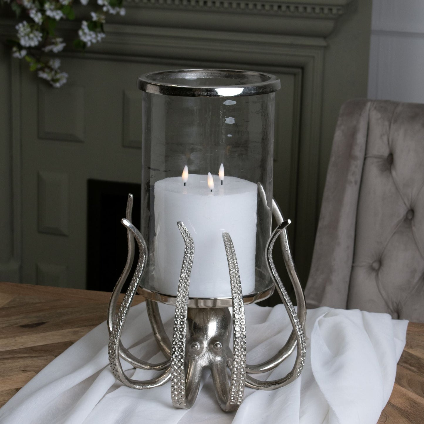 lifestyle image of large octopus candle holder, with glass hurricane lamp displayed with triple wick white LED candle on wooden table, a matte green fireplace can be seen in the background