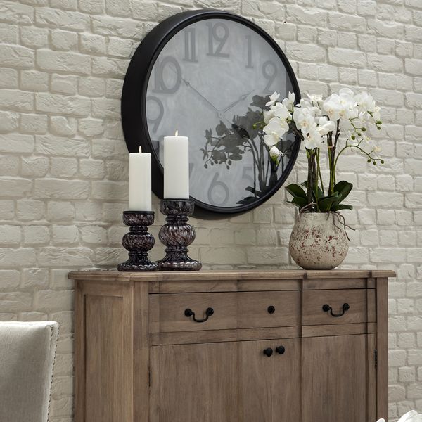 Artificial orchid in stone pot, displayed to the left of a wooden sideboard, with two candlesticks to the left. A large clock is on the wall behind