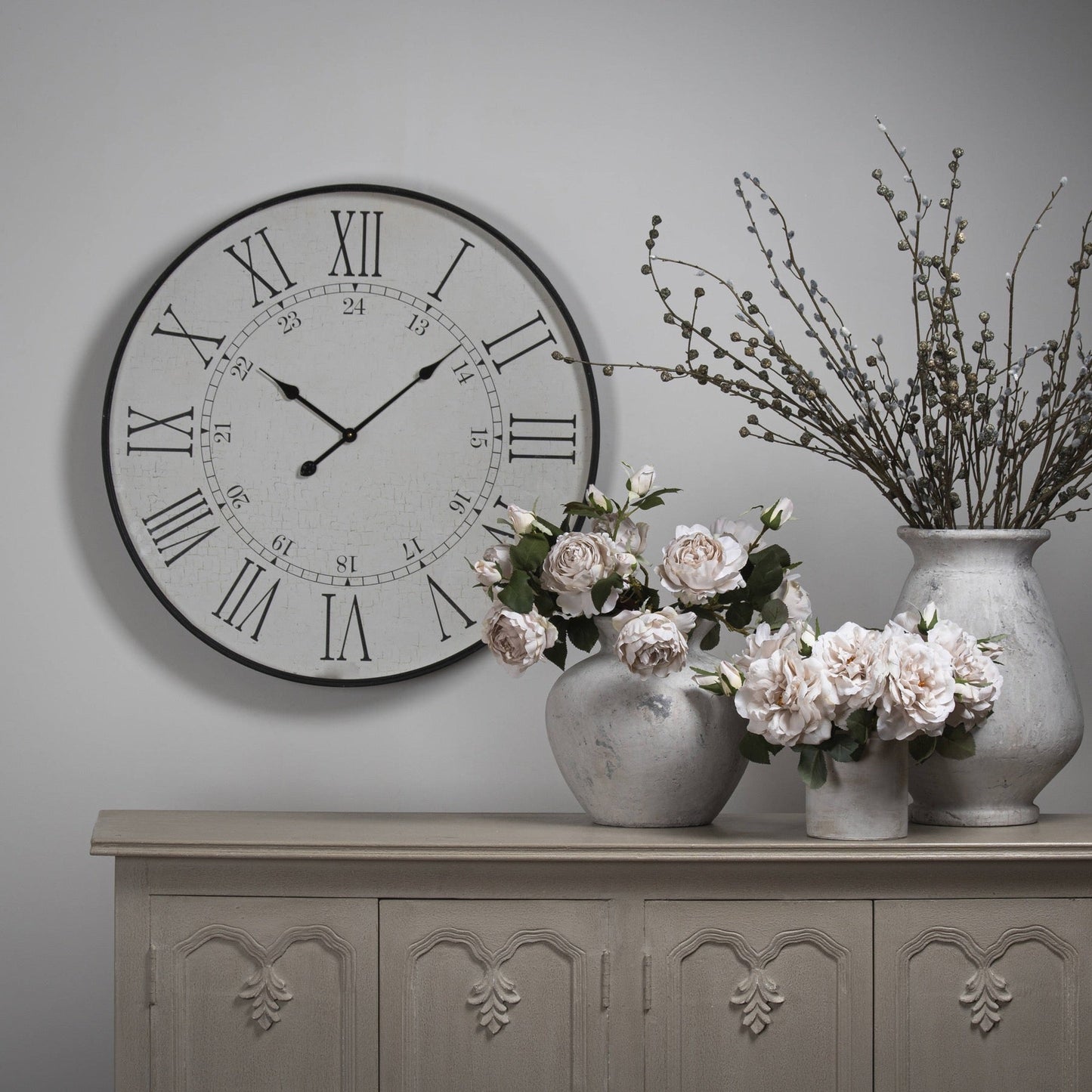 Large station clock mounted on white wall above white washed sideboard. On the sideboard are three varying size stone vases with pale pink artificial rose and dried branches in