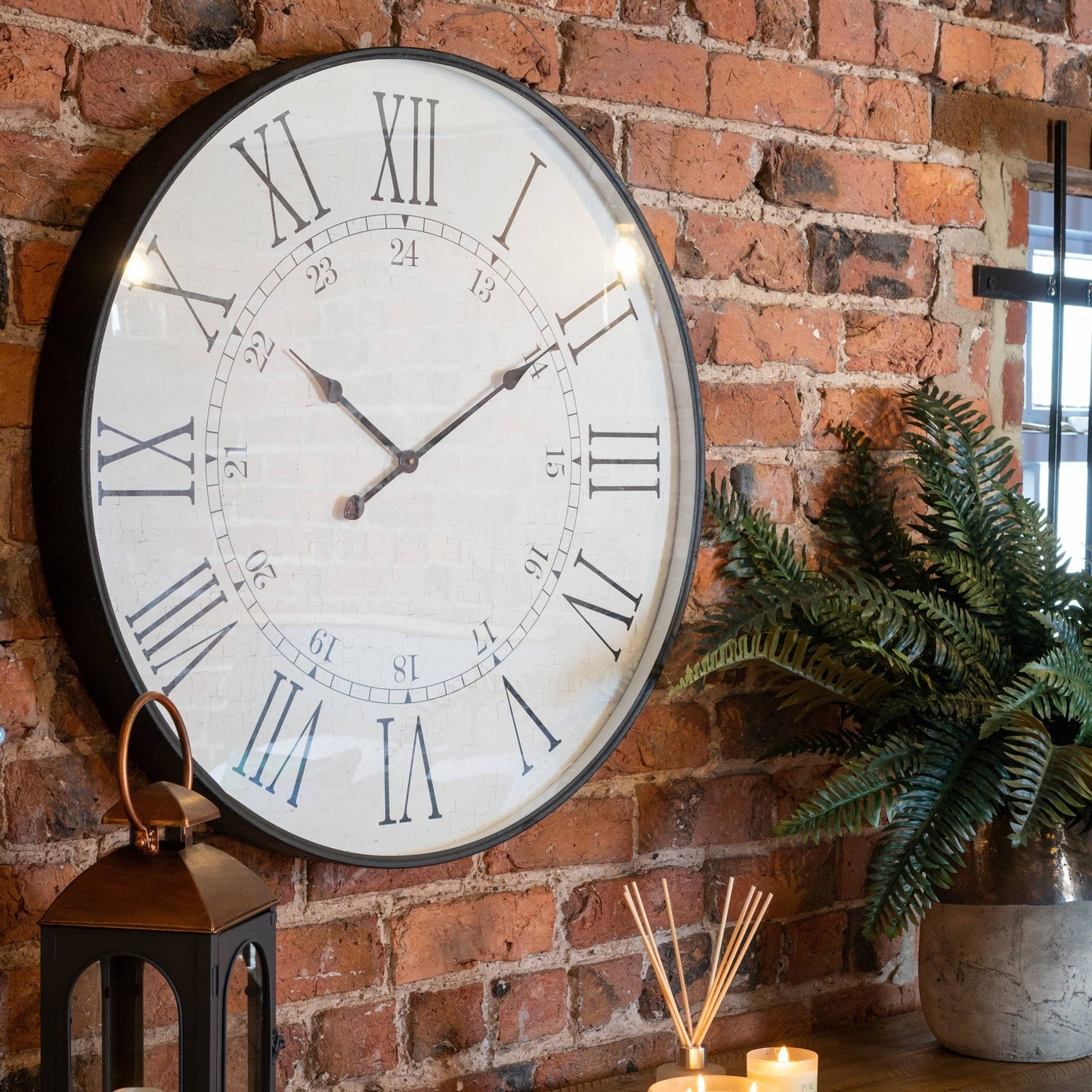 Large station clock wall mounted on exposed brick wall. Artificial fern plant to the right, reed diffuser and candle to the centre and black and bronze lantern to the left