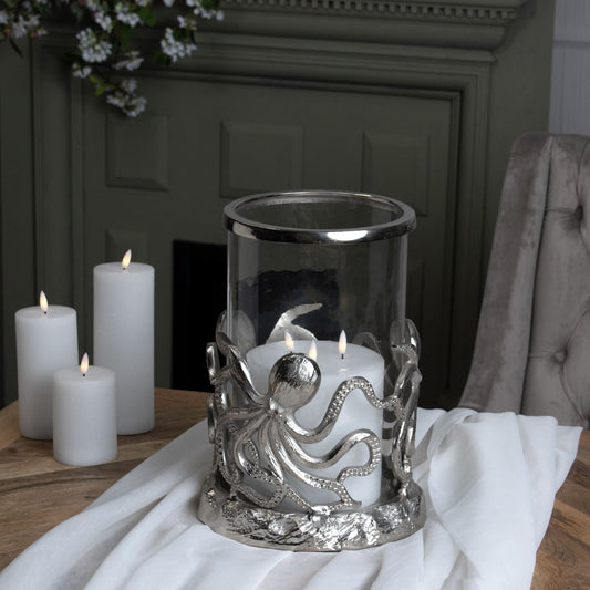 lifestyle image of the Octopus Candle Holder Hurricane Lantern on a wooden top table, with a white linen table cloth draped over the edge. The Lantern stand on top of this with a triple wick white LED candle. Three other smaller size LED Candles are to the left of the lantern on the table and a matte green fireplace can be seen in the background