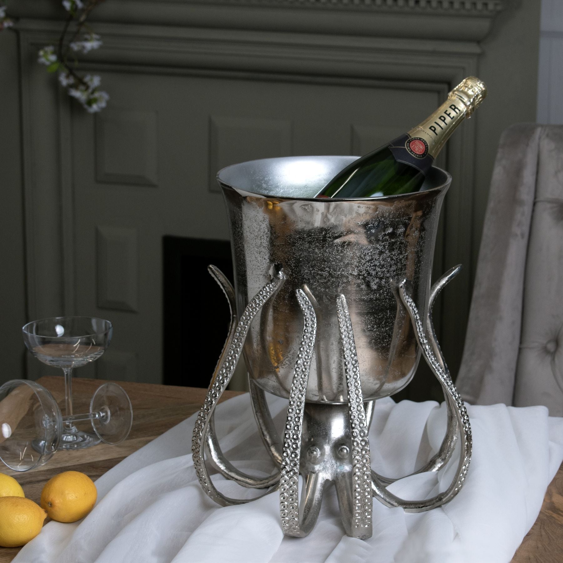 octopus silver champagne bucket on white table cloth next to two champagne coupe glasses and some decorative lemons