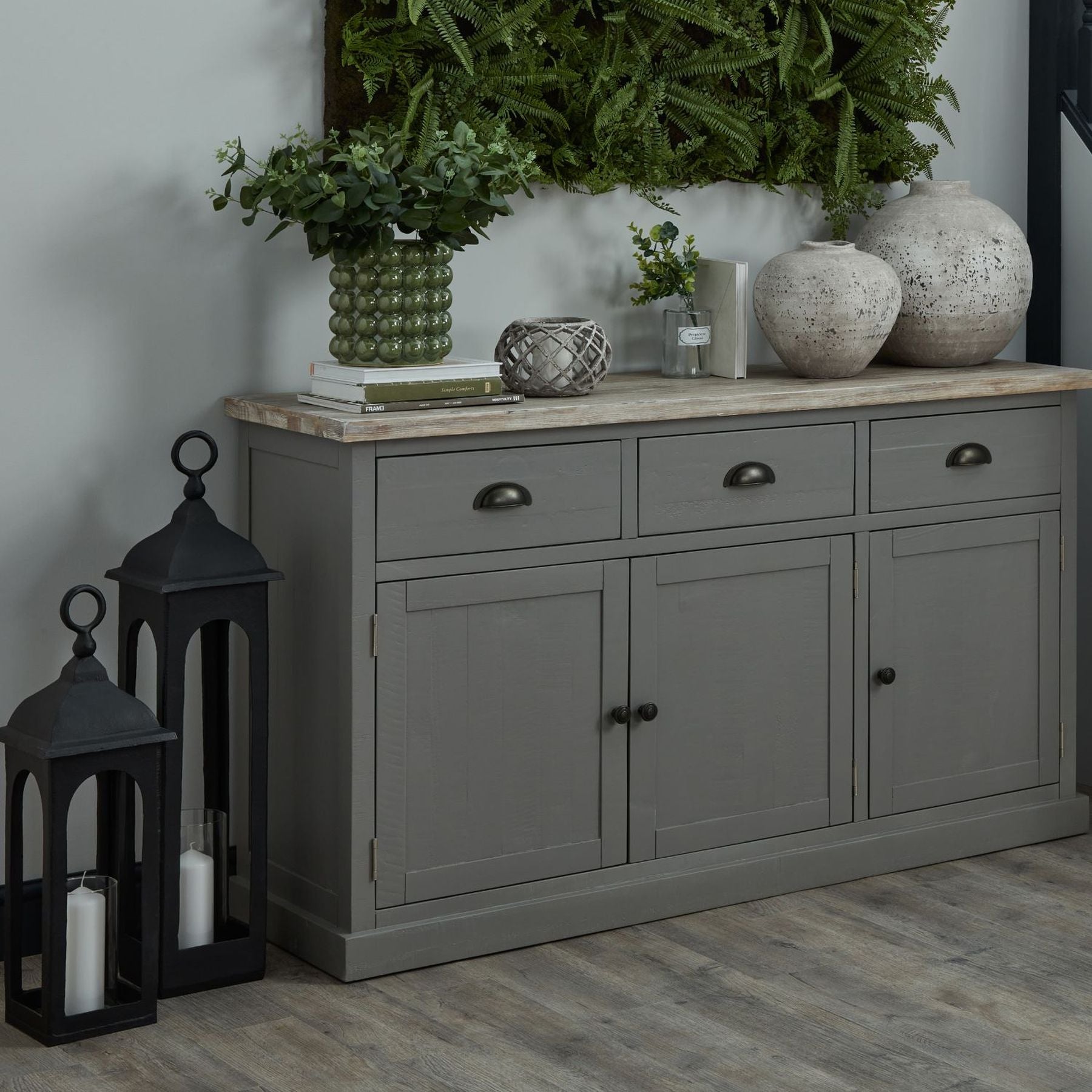 Large Olive green bubble planter on 3 door 3 drawer sideboard, placed on top of some decorative books. An aged stone candle lantern is in the centre, with two varying size aged stone vases to the far side. Two matte black floor standing candle lanterns are on the floor next to the sideboard