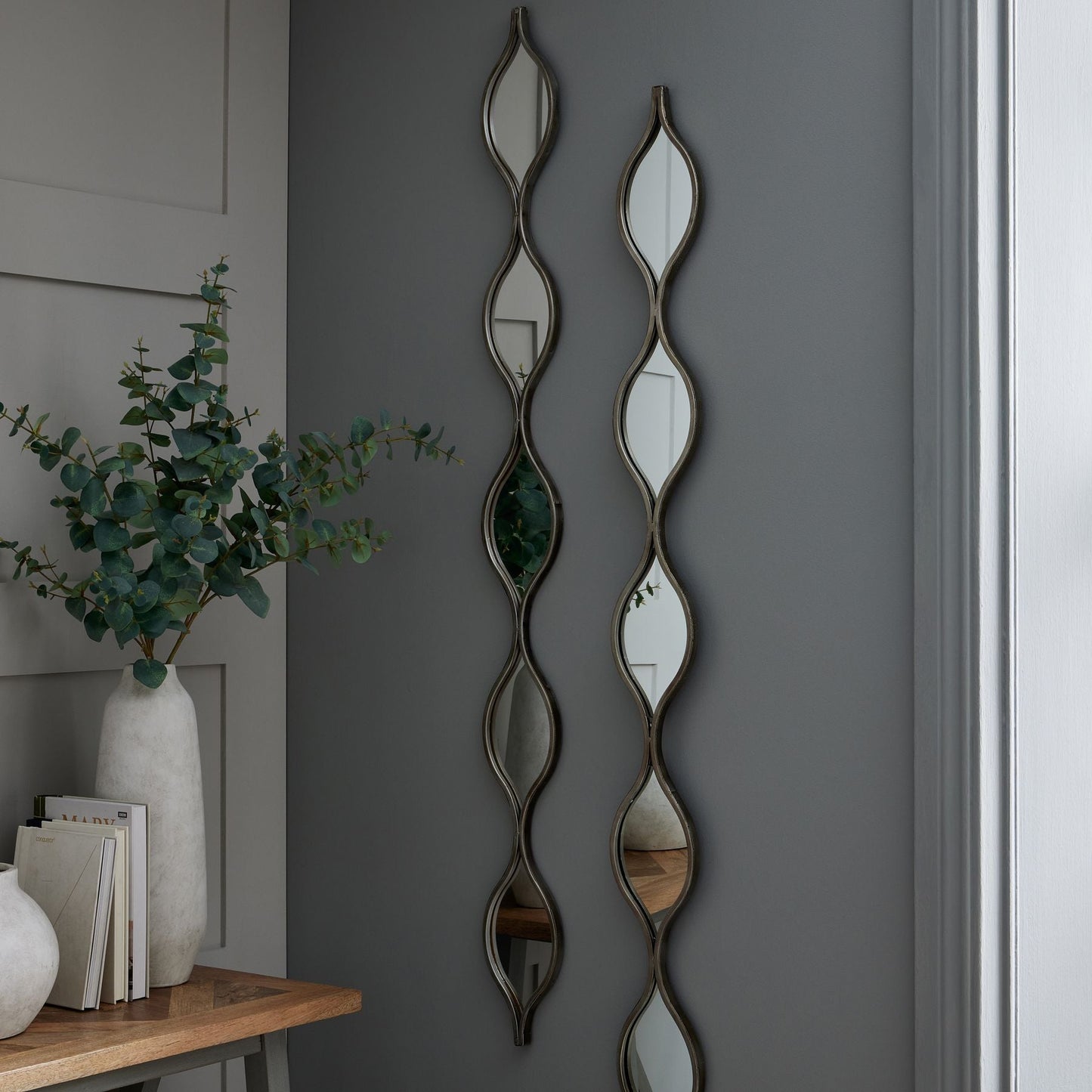 two hanging mirrors on grey wall. Next to console table with stone vase on and artificial eucalyptus stems