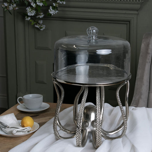 Silver octopus cake stand with glass cloche, on white table cloth, next to small side plate and tea cup and saucer