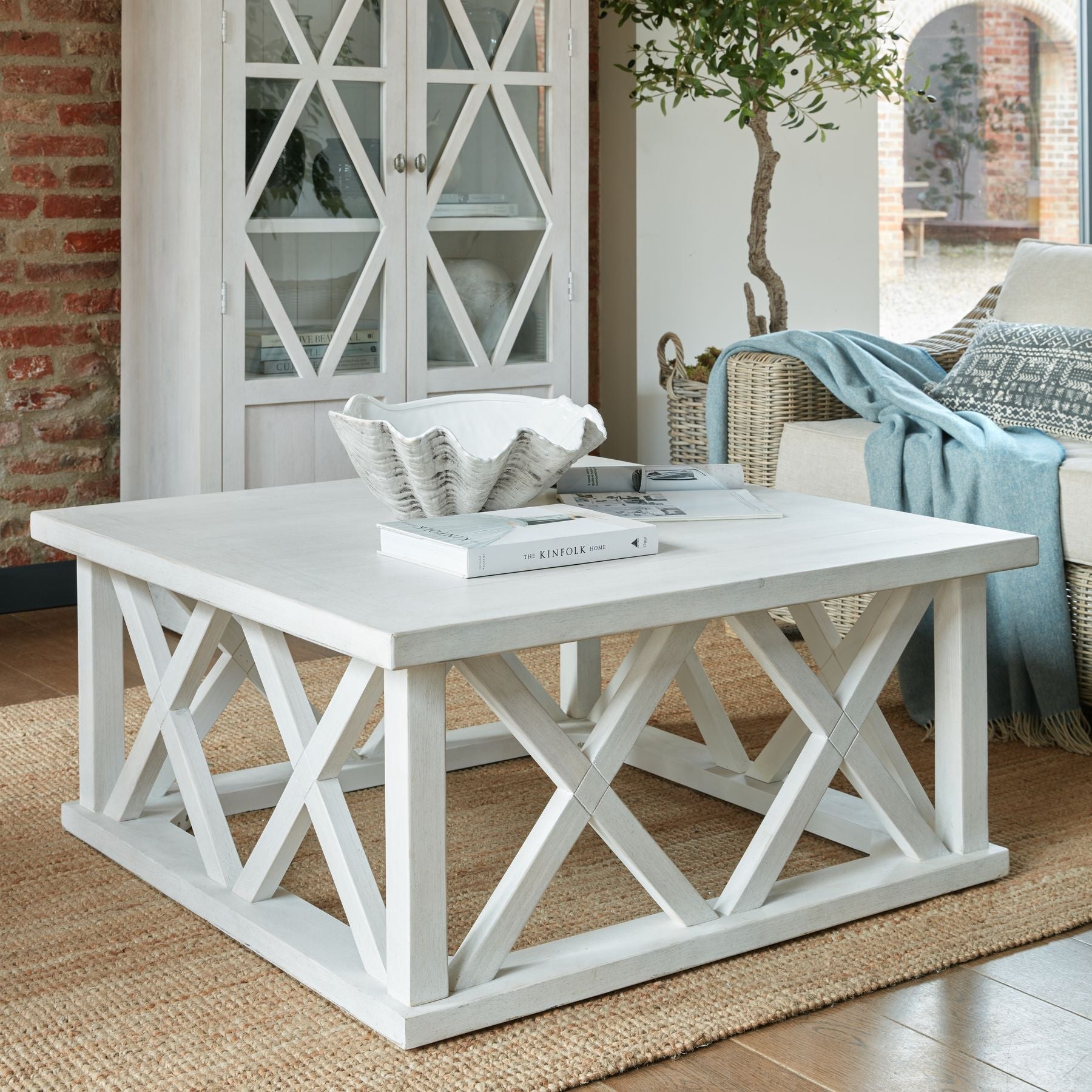 lifestyle image of the square coffee table from the Stanton Furniture Collection in a living room on  a jute rug there is a clamshell vase and decorative books to the top and the matching Stanton Tall Display Cabinet can be seen behind