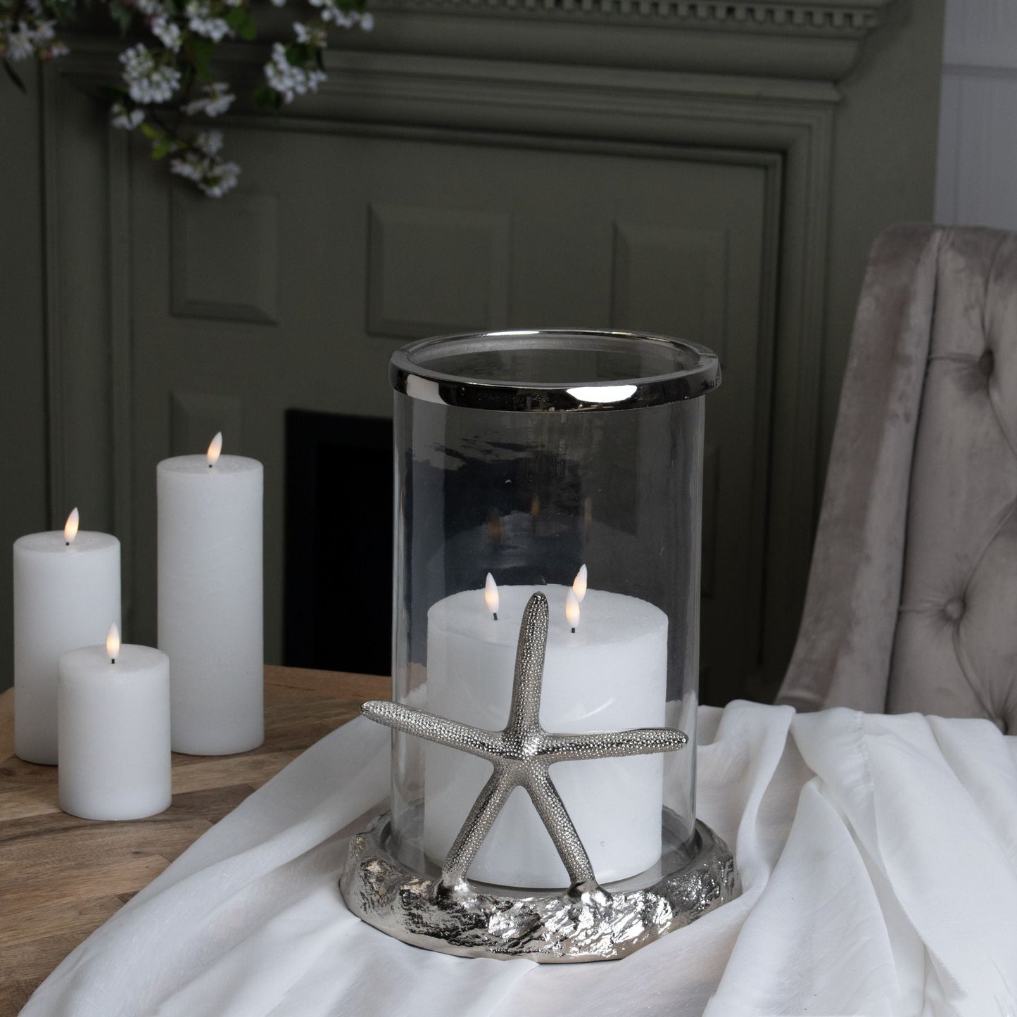 lifestyle image with the hurricane lantern on a table that is covered with white linen. Three LED Candles can be seen to the left of the image and an olive green fireplace is in the background