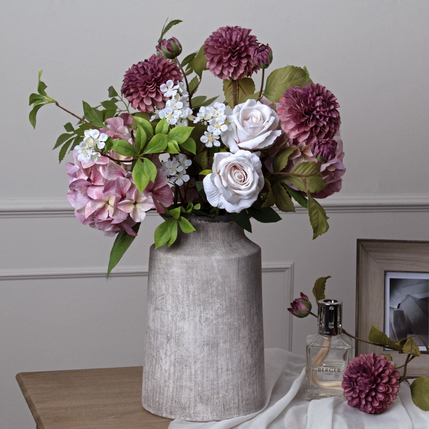 lifestyle image of Urn Vase with forever florals in, on sideboard next to a bottle of perfume, wood effect photo frame and laid down artificial dahlia