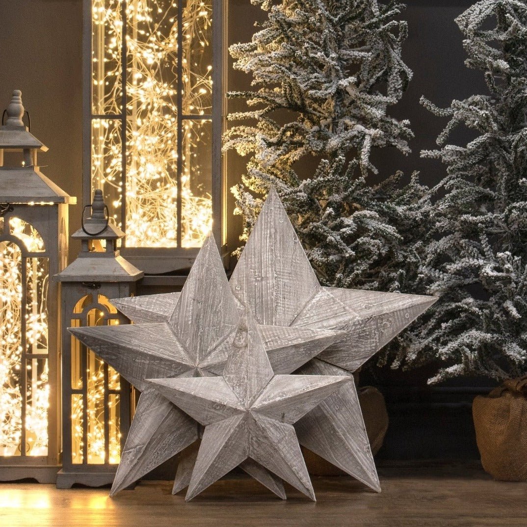 lifestyle image on the three wooden stars freestanding on a wooden floor. Frosted christmas trees can be seen to the background on the right side and lanterns with micro string lights to the left